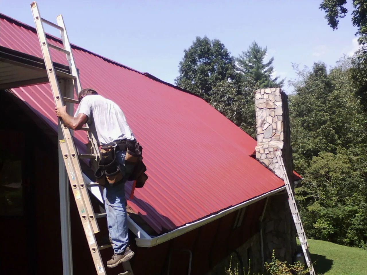 Roofer on a ladder beside a red metal roof and stone chimney — Soffit & Fascia Repair by Roofing Brothers in Westville